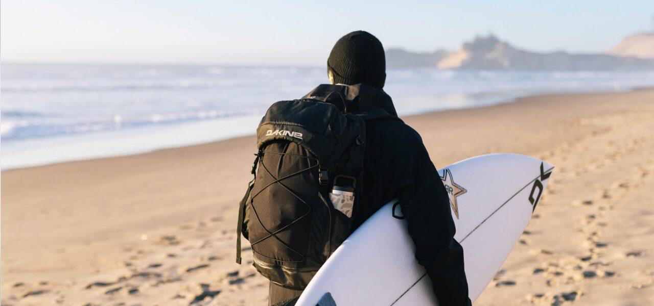 Surfer mit Dakine-Rucksack und Surboard unter dem Arm